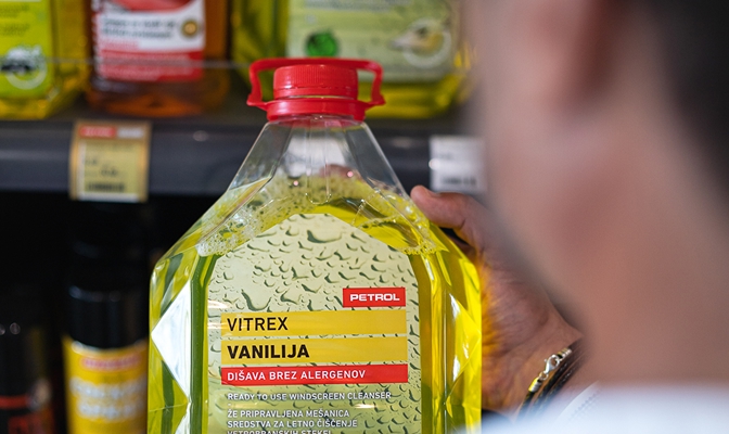 Na sliki je plastenka tekočine za čiščenje vetrobranskega stekla Petrol Vitrex z vonjem vanilje, ki jo oseba drži v roki pred prodajno polico z izdelki za avtokozmetiko.
