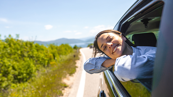 Otrok se naslonjen skozi odprto okno avtomobila sproščeno ozira na sončno podeželsko pokrajino ob cesti.
