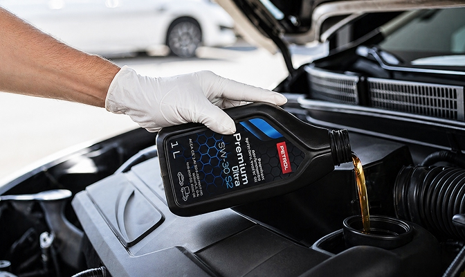 Roka v beli zaščitni rokavici doliva motorno olje Petrol Premium Ultra v motorni prostor avtomobila. Pokrov motorja je odprt, olje pa teče iz črne plastenke v odprtino za dolivanje.