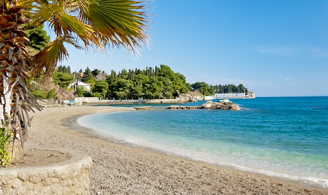 Mirna prodnata plaža ob turkiznem morju, obdana z borovci in sredozemskim rastlinjem. V ospredju so palma in kamnit zid, v ozadju pa obala z belimi stavbami pod jasnim modrim nebom.
