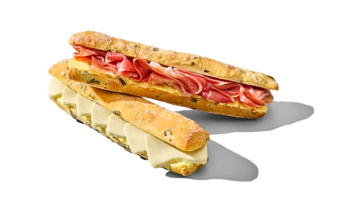 Na sliki sta dva podolgovata sendviča na beli podlagi – eden polnjen s tankimi rezinami pršuta, drugi s svetlim sirom mozzarella, v hrustljavi štručki z olivami.