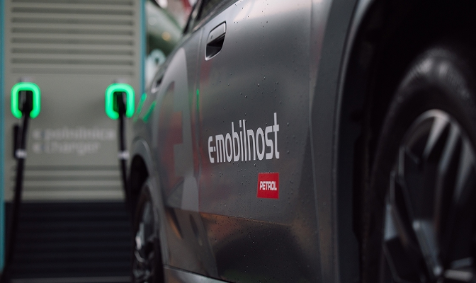 Električno vozilo Petrolove storitve eMobilnost med polnjenjem na električni polnilnici. V ospredju je bočna stran avtomobila z logotipom eMobilnost in Petrol, v ozadju pa sta vidna priključka polnilne postaje z zeleno osvetlitvijo