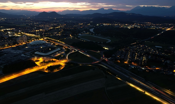 Zračni pogled na osvetljeno cestno in urbano infrastrukturo ob mraku. V ospredju so prometna vozlišča in cestne povezave, v ozadju pa mesto ter gorska panorama pod večernim nebom.