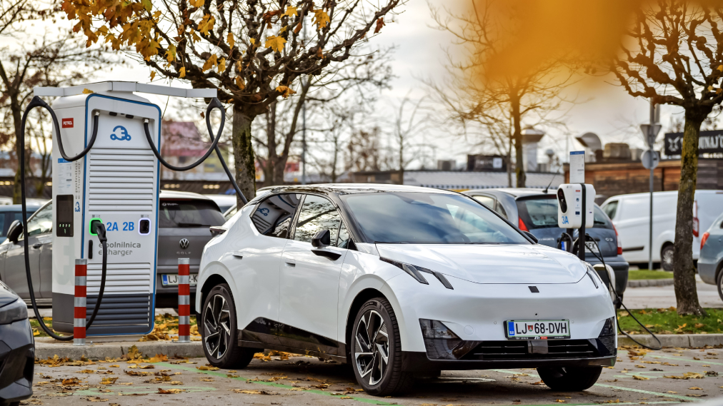 Bel električni avtomobil je priključen na Petrolovo e-polnilnico na zunanjem parkirišču, v ozadju so druga vozila in drevesa z jesenskim listjem.